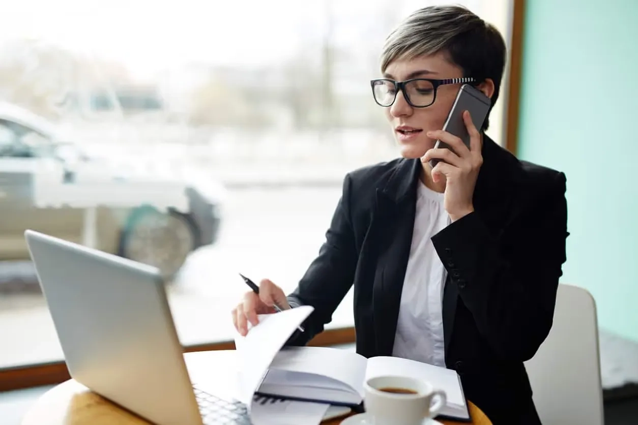 A woman wearing glasses having a phone conversation at a table with a laptop.