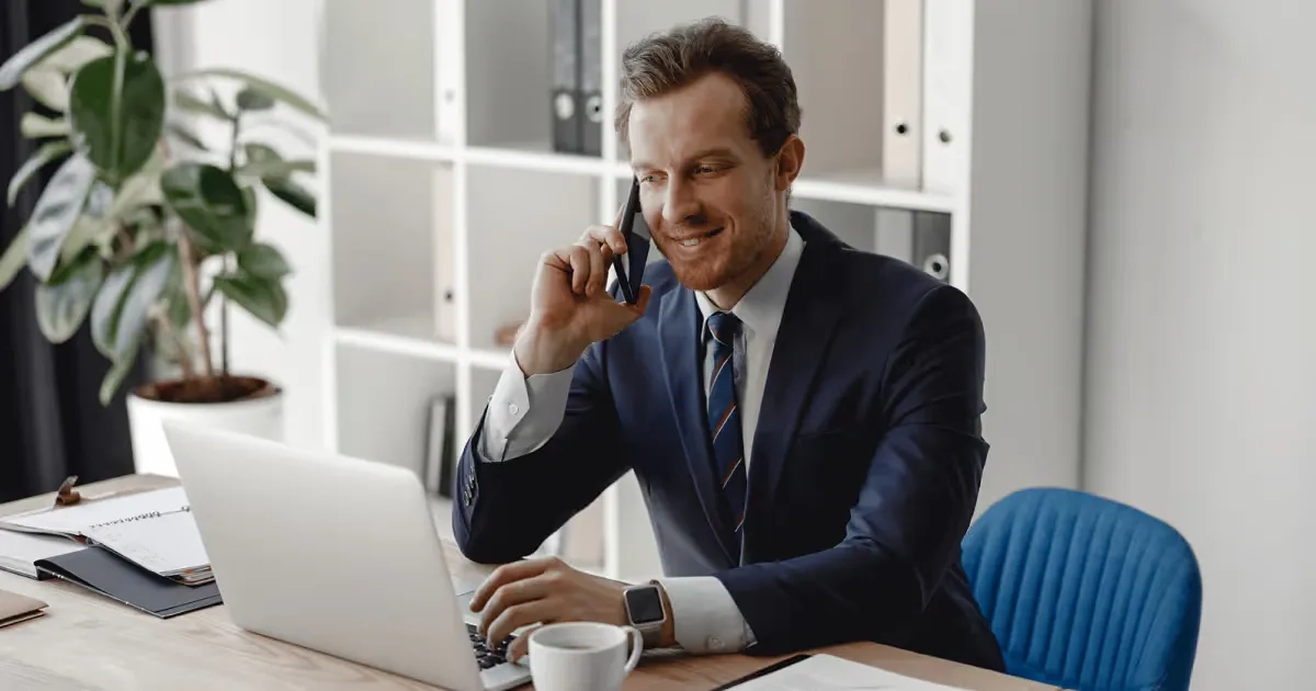 A man in a suit and tie sitting at a desk with a laptop and talking on the phone.