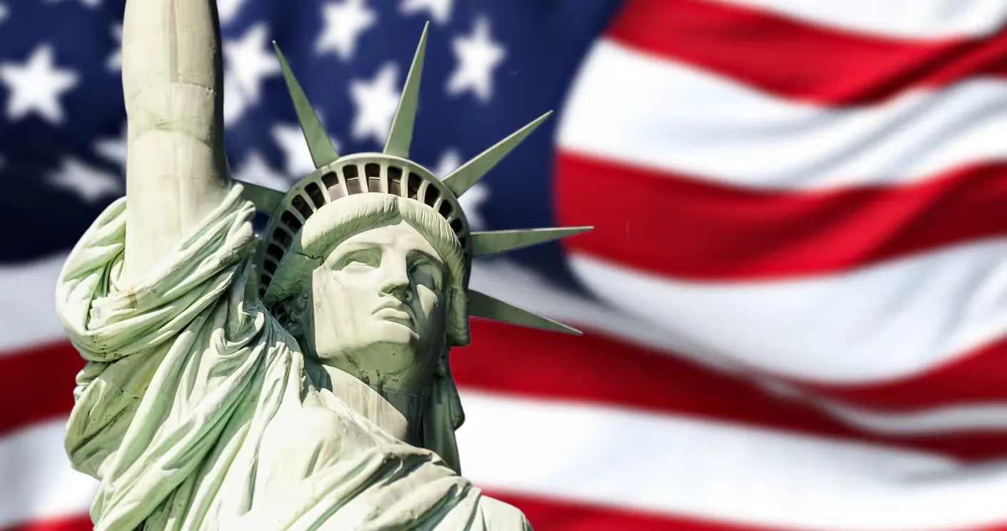 The Statue of Liberty, shown from the shoulders up, stands proudly in front of a rippling American flag, symbolizing the central role of immigration in the 2024 Presidential Election.