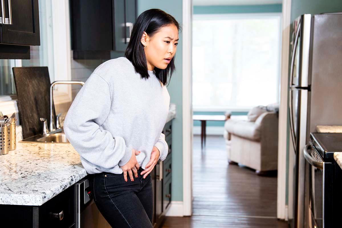A woman holding her abdominal region in pain standing in her kitchen