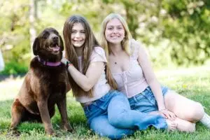 Anna sits in between her older sister and brown lab in the green grass