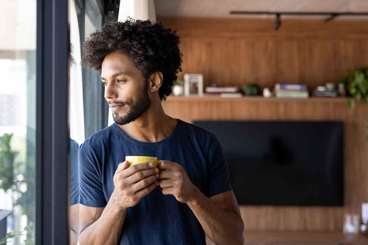 A man looking out the window with a mug of warm water in his hands