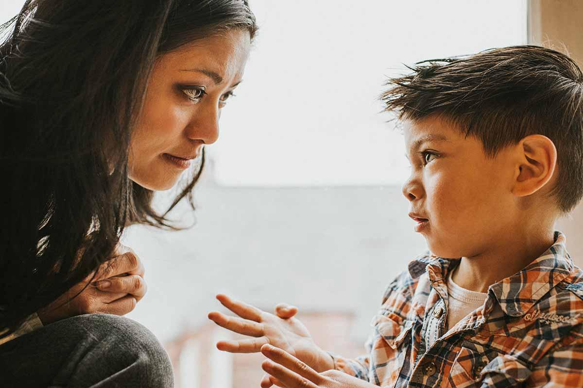A cute young boy gestures using his hands. A woman, who sits close-by, listens and watches him intently as he explains something.