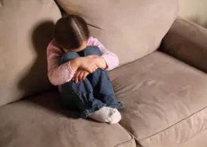 Child hugs knees and hides face while sitting on couch