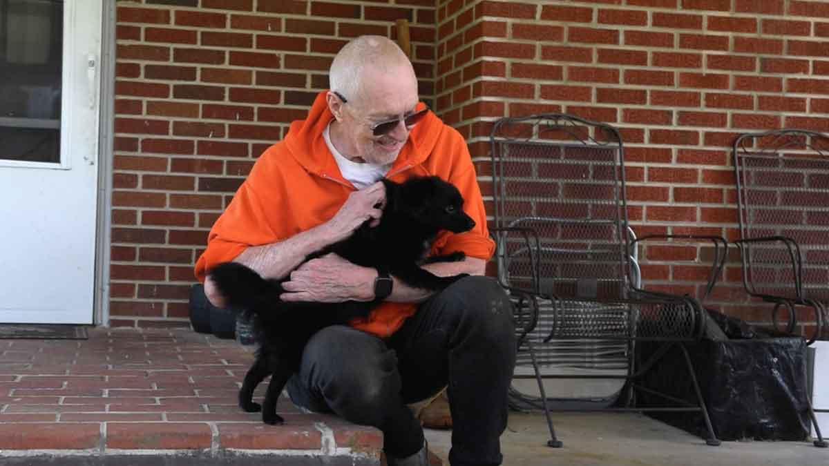 Jerry Austin wearing an orange shirt and dark pants, sitting on a porch and holding a black dog. 
