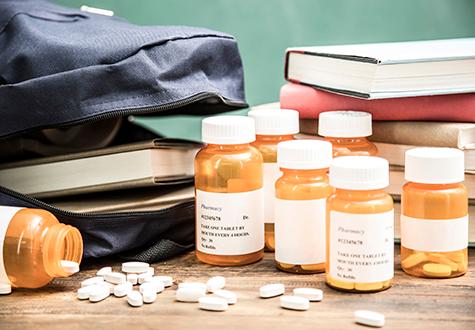 Prescription drug abuse in school concept.  Many pill bottles and drugs on high school, classroom desk with backpack and stack of textbooks.  Chalkboard is background.   Various concepts:  drug trafficking, drug abuse, addiction.