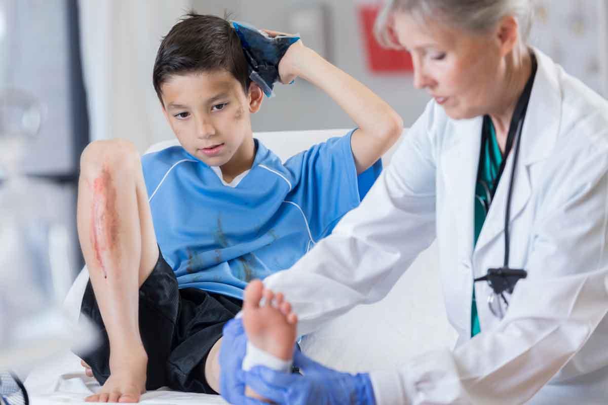 A young boy holding an ice pack to his head, with a cut on his shin and getting his ankle wrapped by a healthcare [rovider